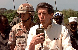 Special Envoy to Somalia, Ambassador Robert Oakley, is seen from the chest up, facing the camera and speaking to a group of Somalis over a public address system. Behind him and to the left is US Army BGEN Lawson W. Magruder III, Vice Commander, 10th Mountain Division (Light), in Somalia as the Commander of Combined Task Force Kismayo. Somali civilians stand in the background. This mission is in direct support of Operation Restore Hope.