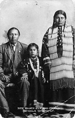 Nicholas Black Elk, daughter Lucy Black Elk and wife Anna Brings White, photographed in their home in Manderson, South Dakota, ca 1910. Black Elk wears a suit, his wife wears a long dress decorated with elk's teeth and a hair pipe necklace. (Source: The Sixth Grandfather, edited by Raymond DeMallie p. 260)