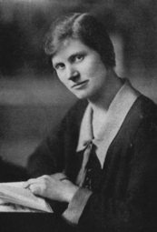 A white woman with side-parted straight dark hair, wearing a collared dress, hand on a book